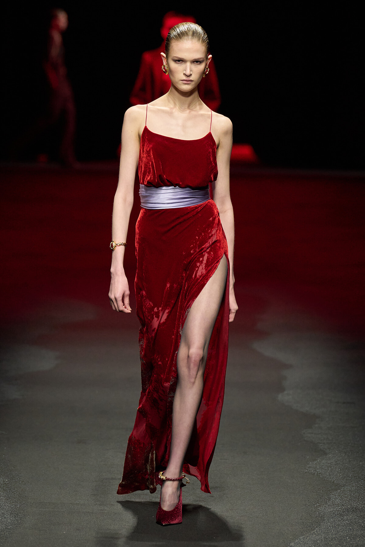 Model walking the runway in a red velvet slip dress with a silver sash and high slit.