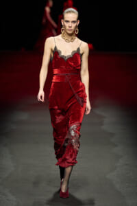 Model walking the runway in a red velvet slip dress with black lace trim and a tasselled belt, wearing bold gold jewelry (necklace and earrings).