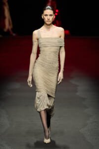 Model walking a runway in a beige ruched off-shoulder midi dress with long matching gloves and ballet heels.