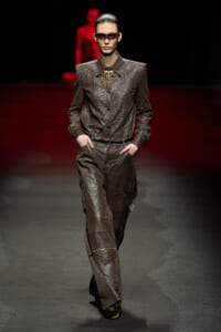 Fashion model walking a runway in a textured brown suit with a wide collar and matching cargo pants, hands in pockets, wearing dark sunglasses and an amber-toned statement necklace.