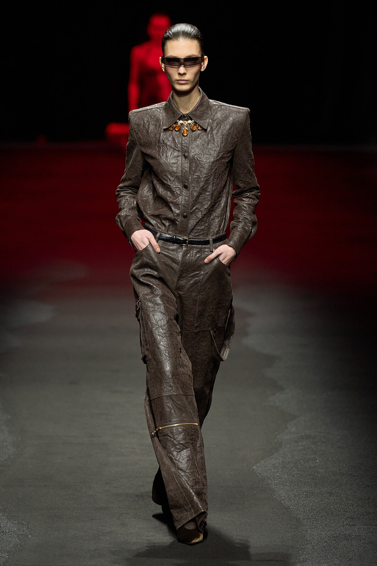 Fashion model walking a runway in a textured brown suit with a wide collar and matching cargo pants, hands in pockets, wearing dark sunglasses and an amber-toned statement necklace.
