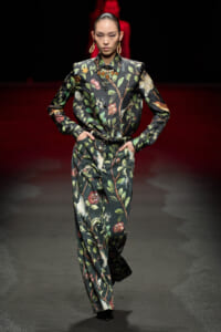 Female model walks a fashion runway in a dark floral suit with a matching jacket and trousers, accessorized with gold earrings and a bold necklace.