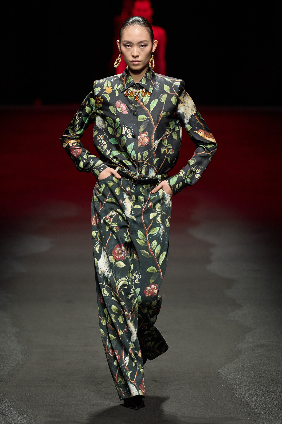Female model walks a fashion runway in a dark floral suit with a matching jacket and trousers, accessorized with gold earrings and a bold necklace.