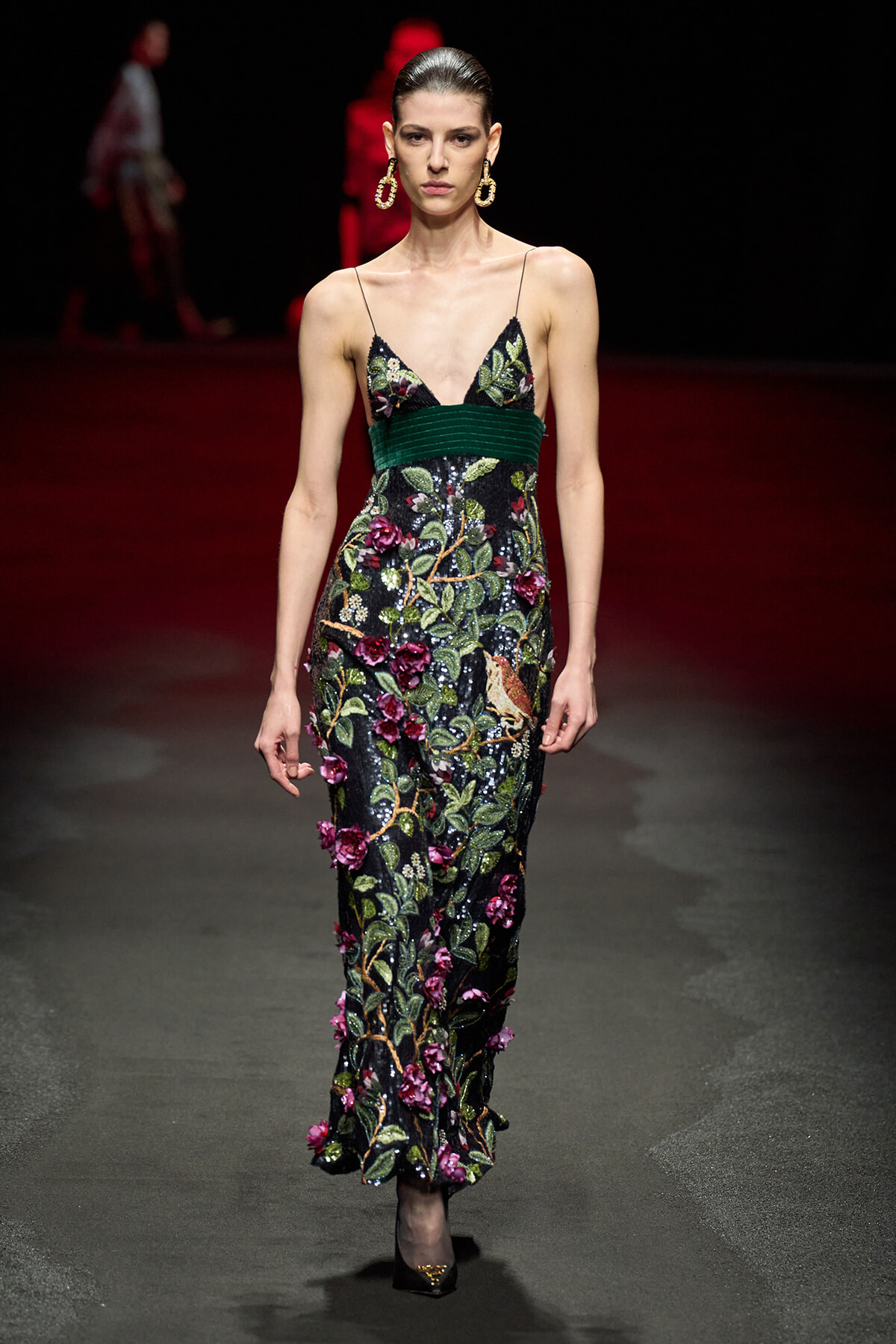 Model walks the runway in a black sequined gown with vibrant floral embroidery and a green waist sash, paired with gold hoop earrings.