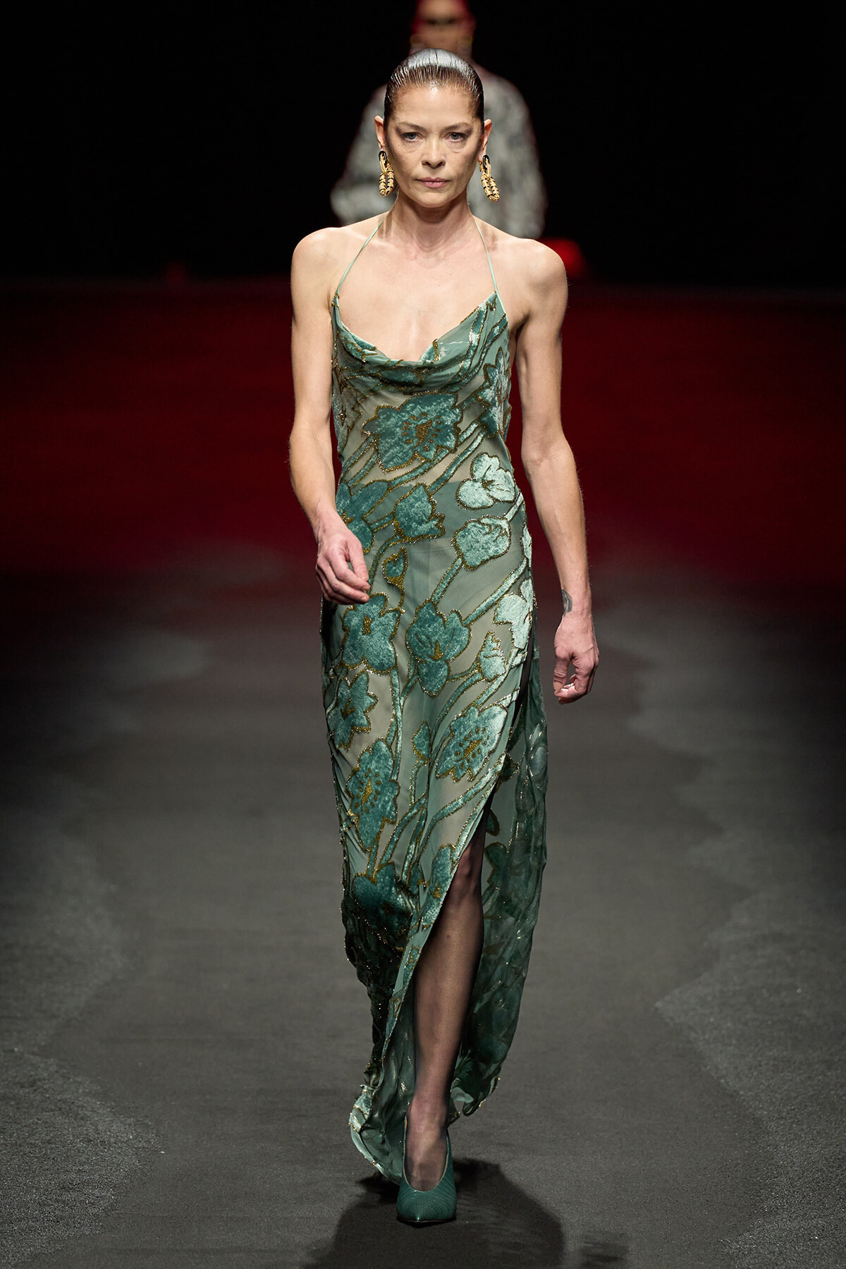Female model walking a runway in a green floral evening gown with a thigh-high slit and matching green heels. large gold earrings stand out.