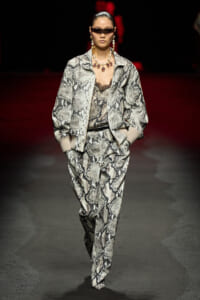 Model walks a snakeskin-patterned suit on a runway, wearing oversized jacket, matching pants, sunglasses, and bold jewelry.
