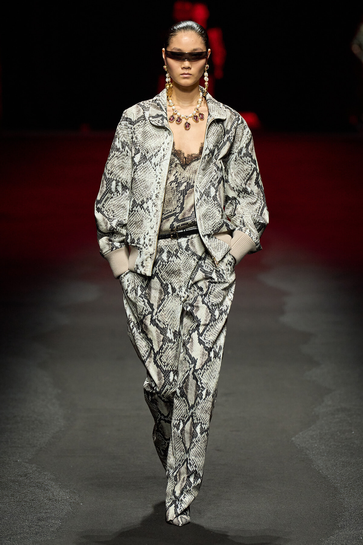 Model walks a snakeskin-patterned suit on a runway, wearing oversized jacket, matching pants, sunglasses, and bold jewelry.