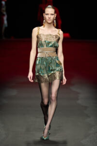 Model walking a fashion runway in a teal-green lace mini dress with gold embroidery and a beige belt, paired with sheer black tights and teal heels.