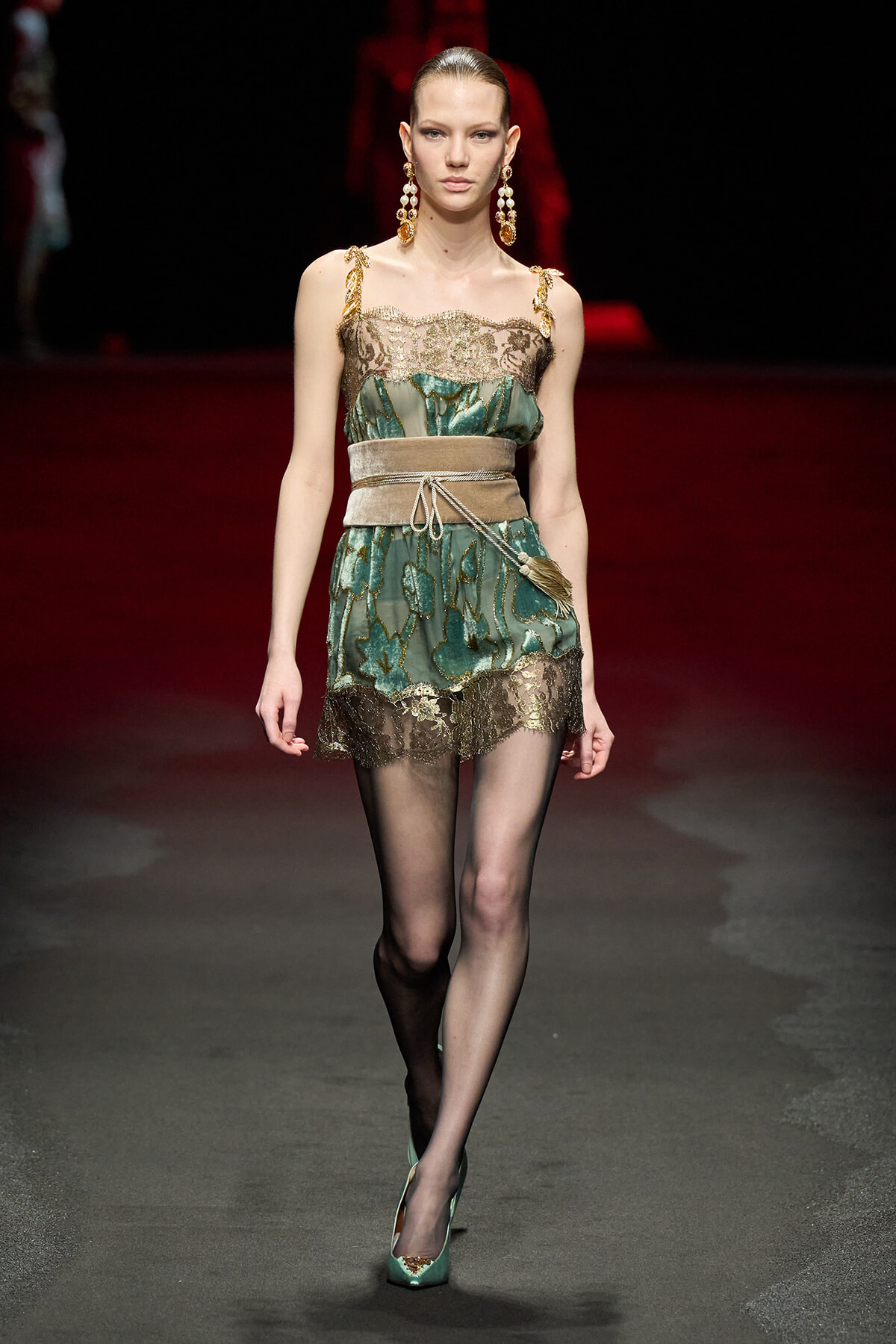 Model walking a fashion runway in a teal-green lace mini dress with gold embroidery and a beige belt, paired with sheer black tights and teal heels.