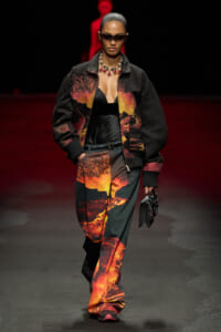 Model walking a runway in a lava-print black bomber jacket and matching pants, black top, statement necklace, futuristic sunglasses, and a small black clutch.