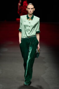 Model on a runway wearing a mint satin button-down blouse and emerald velvet pants, with a gold necklace and green clutch in hand.