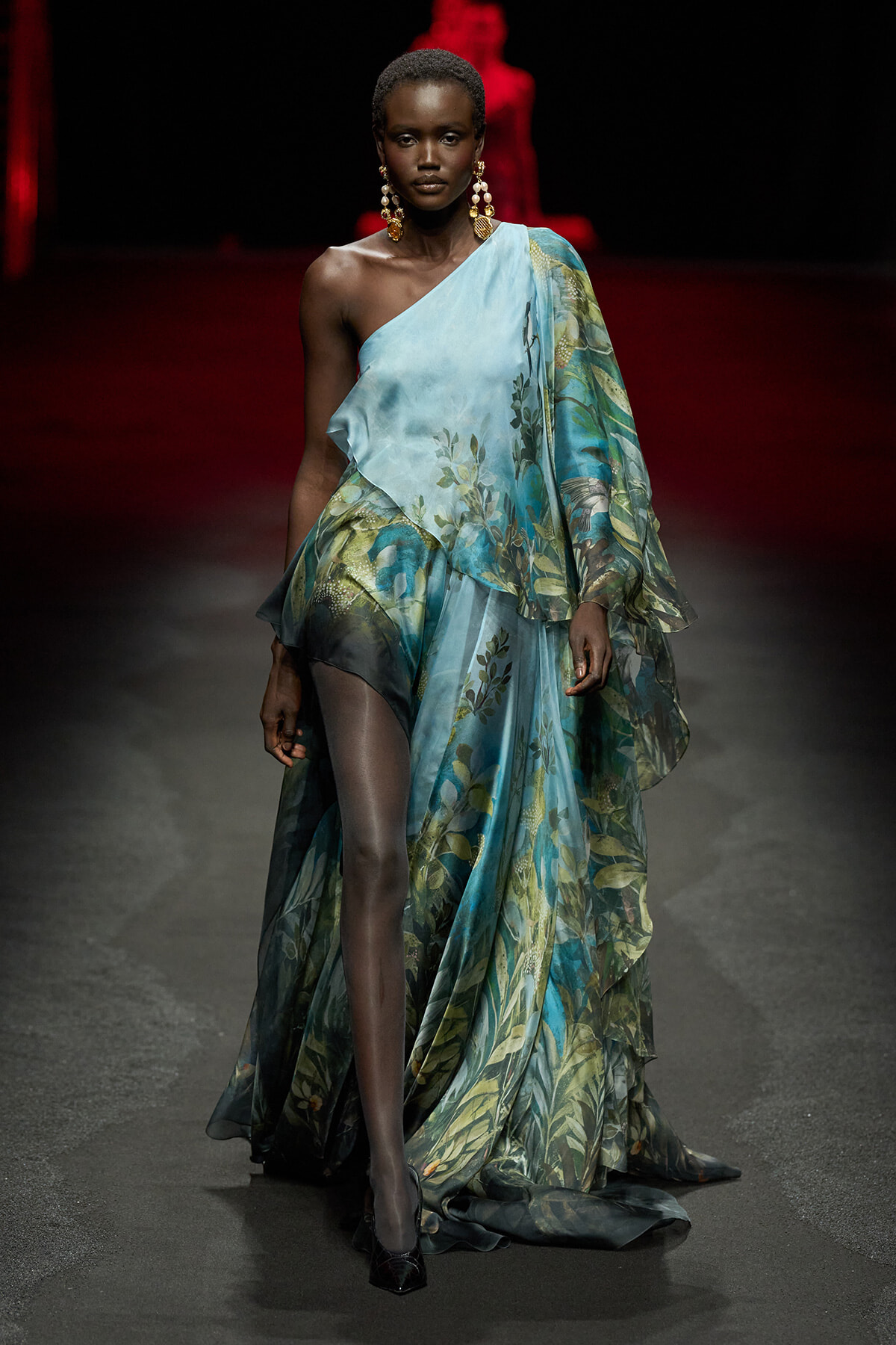 Model wearing a one-shoulder, teal floral gown with sheer layers walking a runway.