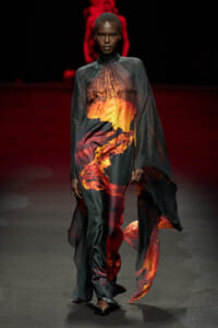 Model walking the runway in a flowing dark gown with a lava-inspired fiery print on the front and cape-like drape.