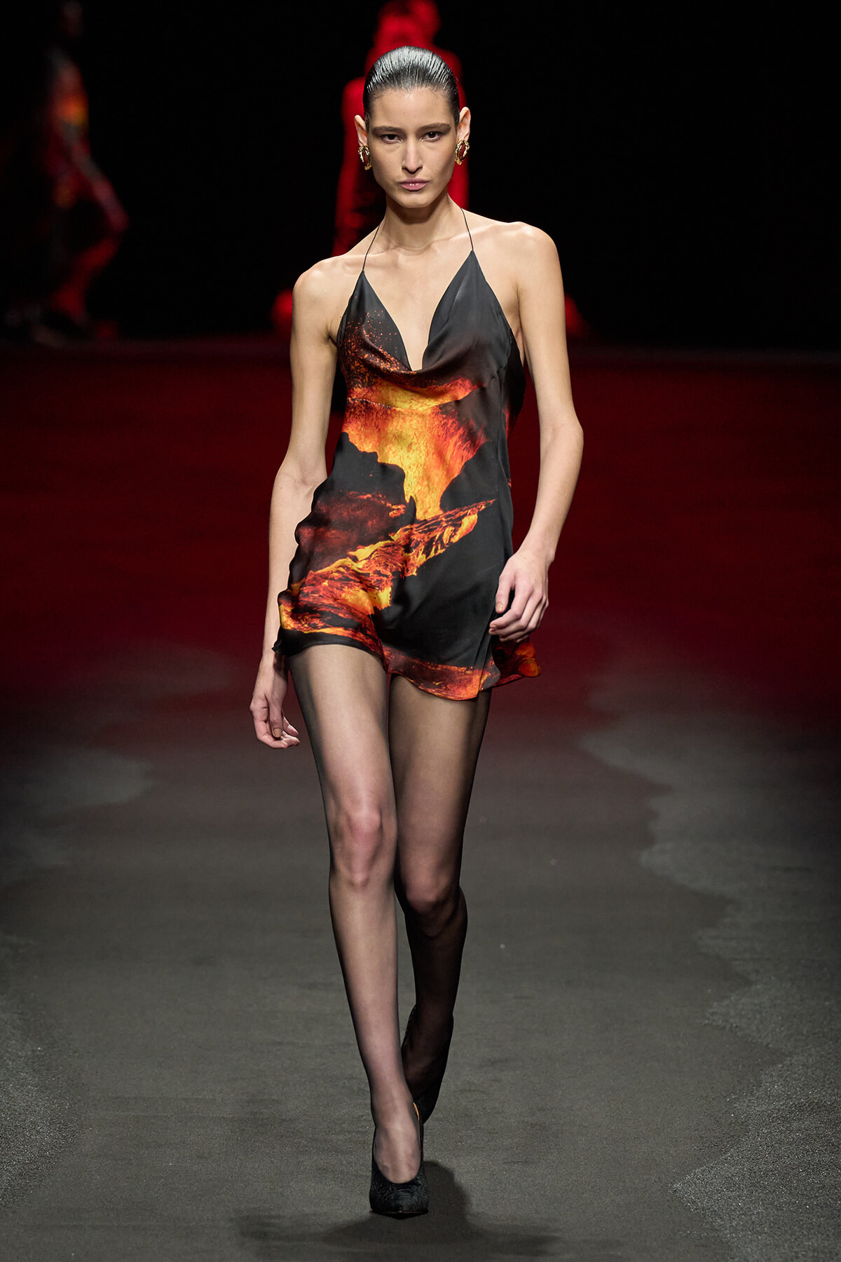 Model on a fashion runway wearing a black lava-print halter romper with fiery orange-red patterns, paired with sheer black tights and black heels.