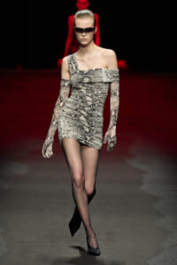 Model walking a fashion runway in a snakeskin-print, one-shoulder mini dress with long matching gloves and dark rectangular sunglasses.