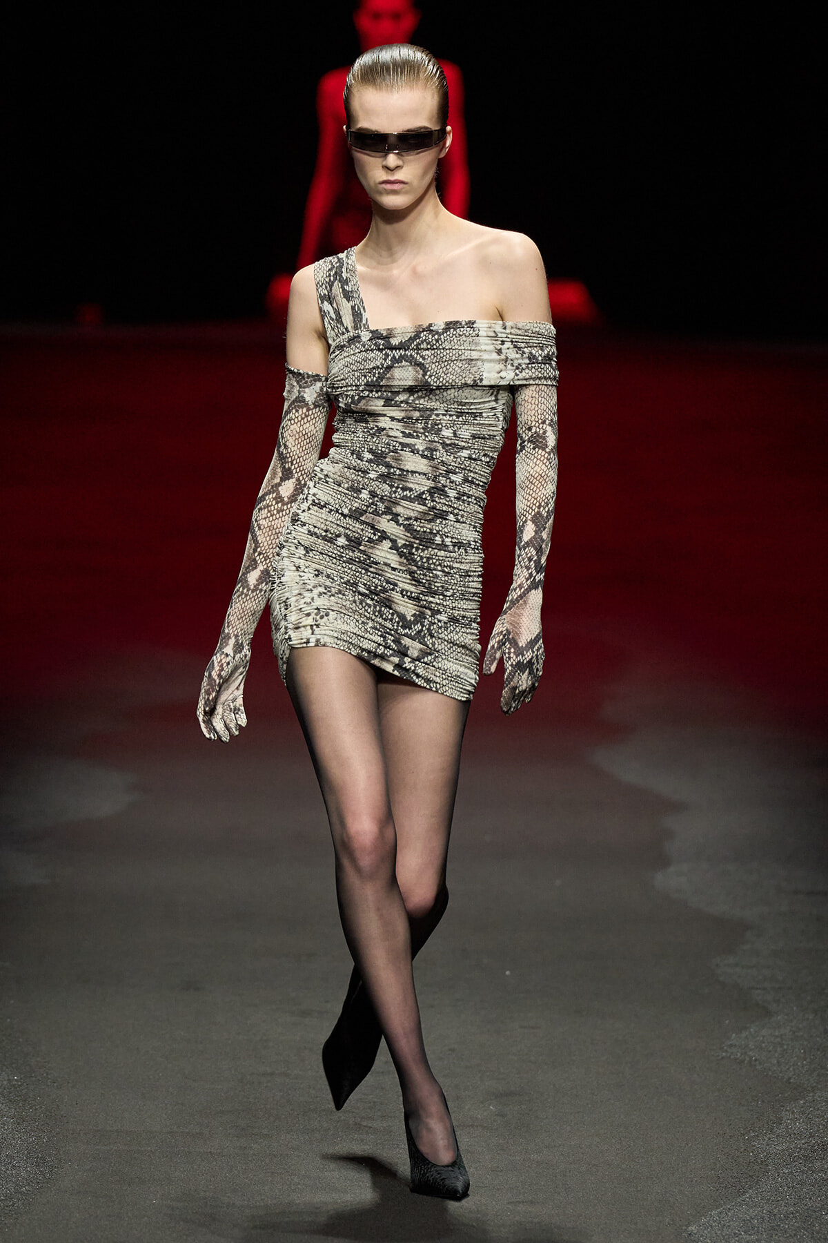 Model walking a fashion runway in a snakeskin-print, one-shoulder mini dress with long matching gloves and dark rectangular sunglasses.