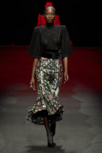 Model walking a runway in a black sheer top with cape sleeves and a high-waisted floral midi skirt.