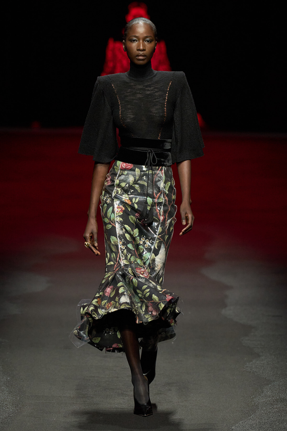 Model walking a runway in a black sheer top with cape sleeves and a high-waisted floral midi skirt.