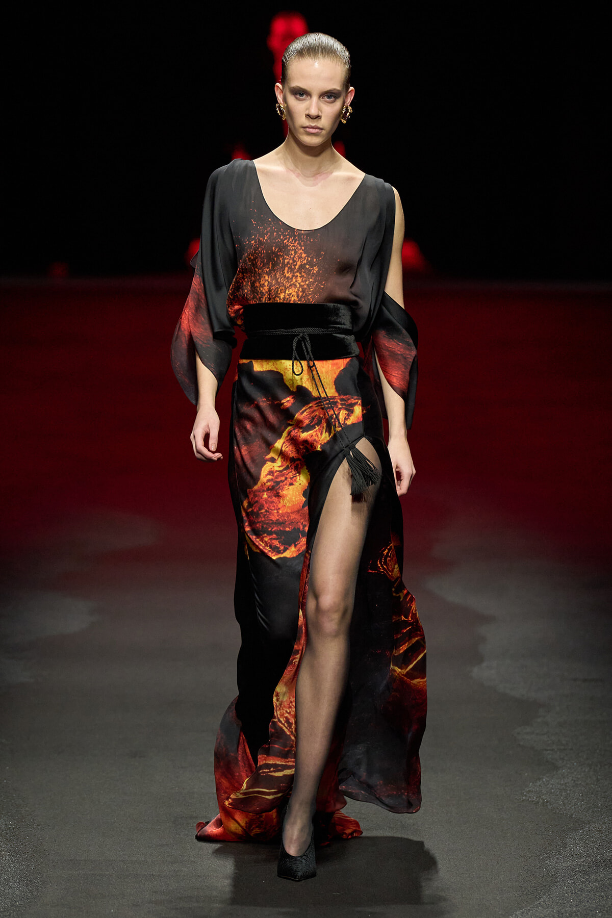 Model walking the runway in a black lava-print gown with a high slit, cinched waist, and dramatic flowing sleeves.