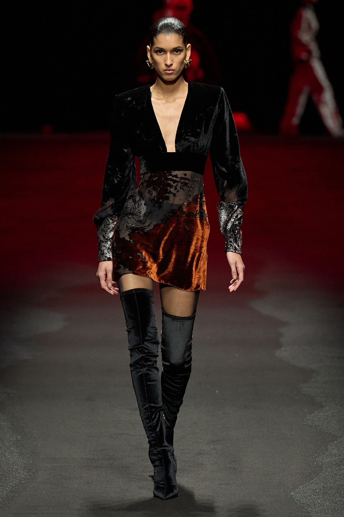 Model walking the runway in a black velvet plunging-neck blouse with a burnt-orange velvet mini skirt and black over-the-knee boots.