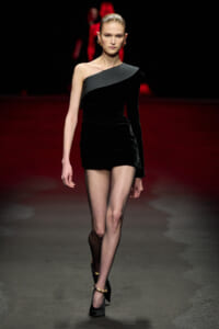 Runway model in a sleek black one-shoulder mini dress, straight hair, and gold earrings walking down a dimly lit catwalk.