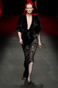 Model walking a runway in a black velvet gown with a deep plunging neckline and lace-appliqué skirt.