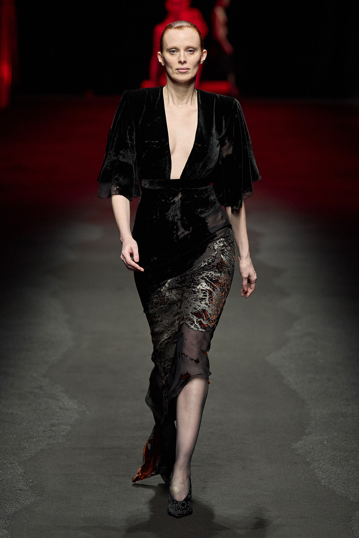 Model walking a runway in a black velvet gown with a deep plunging neckline and lace-appliqué skirt.