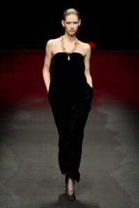 Model on a runway wearing a black strapless velvet jumpsuit with a gold statement necklace, hands in pockets, facing the camera.