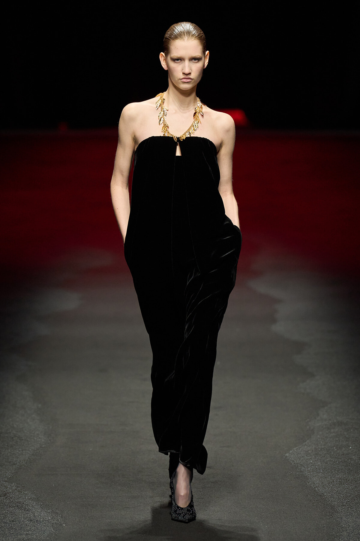 Model on a runway wearing a black strapless velvet jumpsuit with a gold statement necklace, hands in pockets, facing the camera.