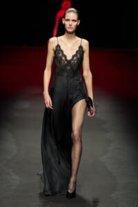Model walking a fashion show in a black satin slip dress with lace bodice and a high thigh slit.