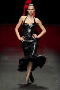 Model walking a runway in a black patent-leather halter-neck mermaid gown, hands on hips.