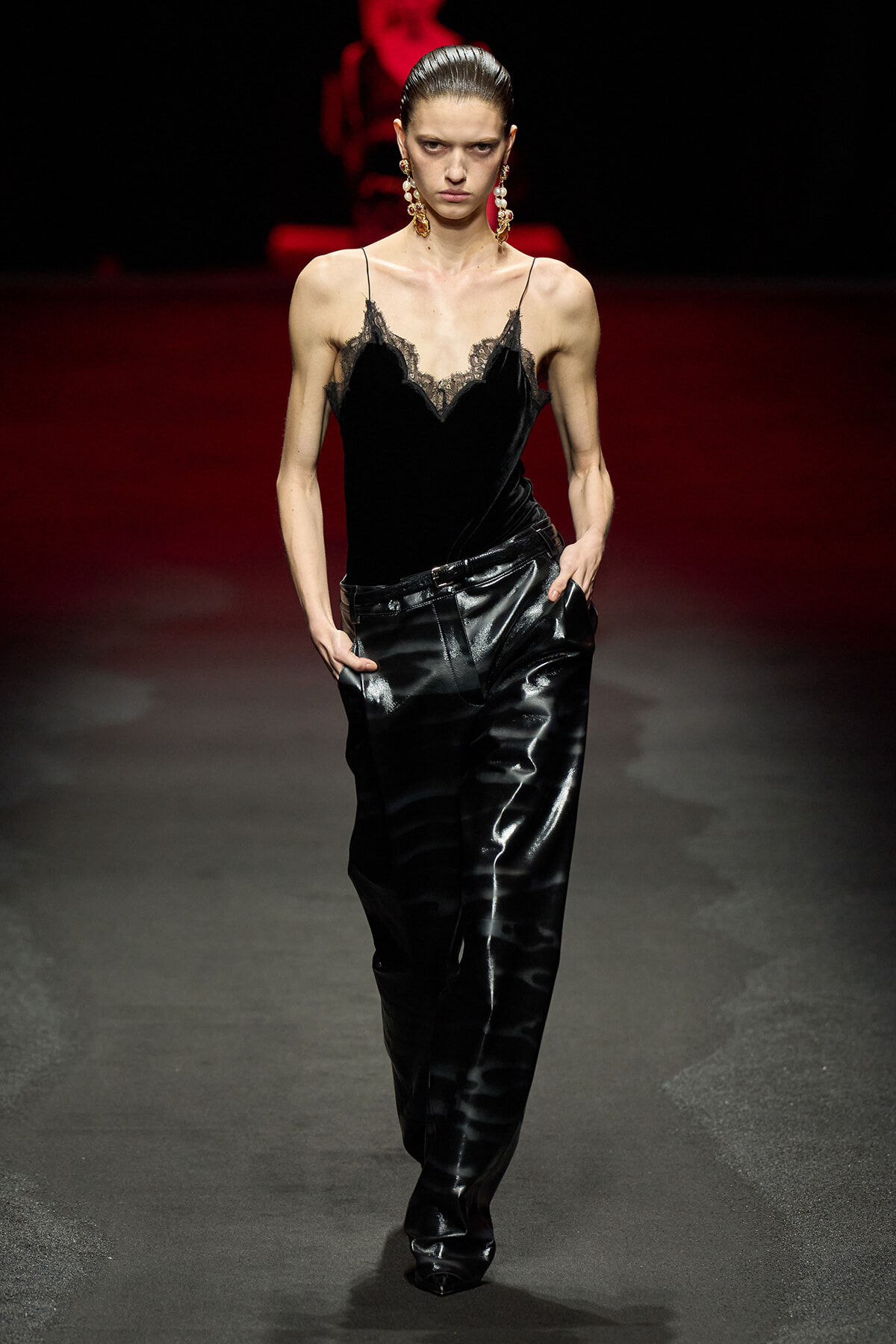 Model walks a fashion runway wearing a black velvet camisole with lace trim and high-shine black leather pants, accessorized with statement dangling earrings.