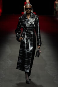 Model wearing a glossy black trench coat with a belt and oversized collar, walking a runway in dark sunglasses while holding a small black handbag.