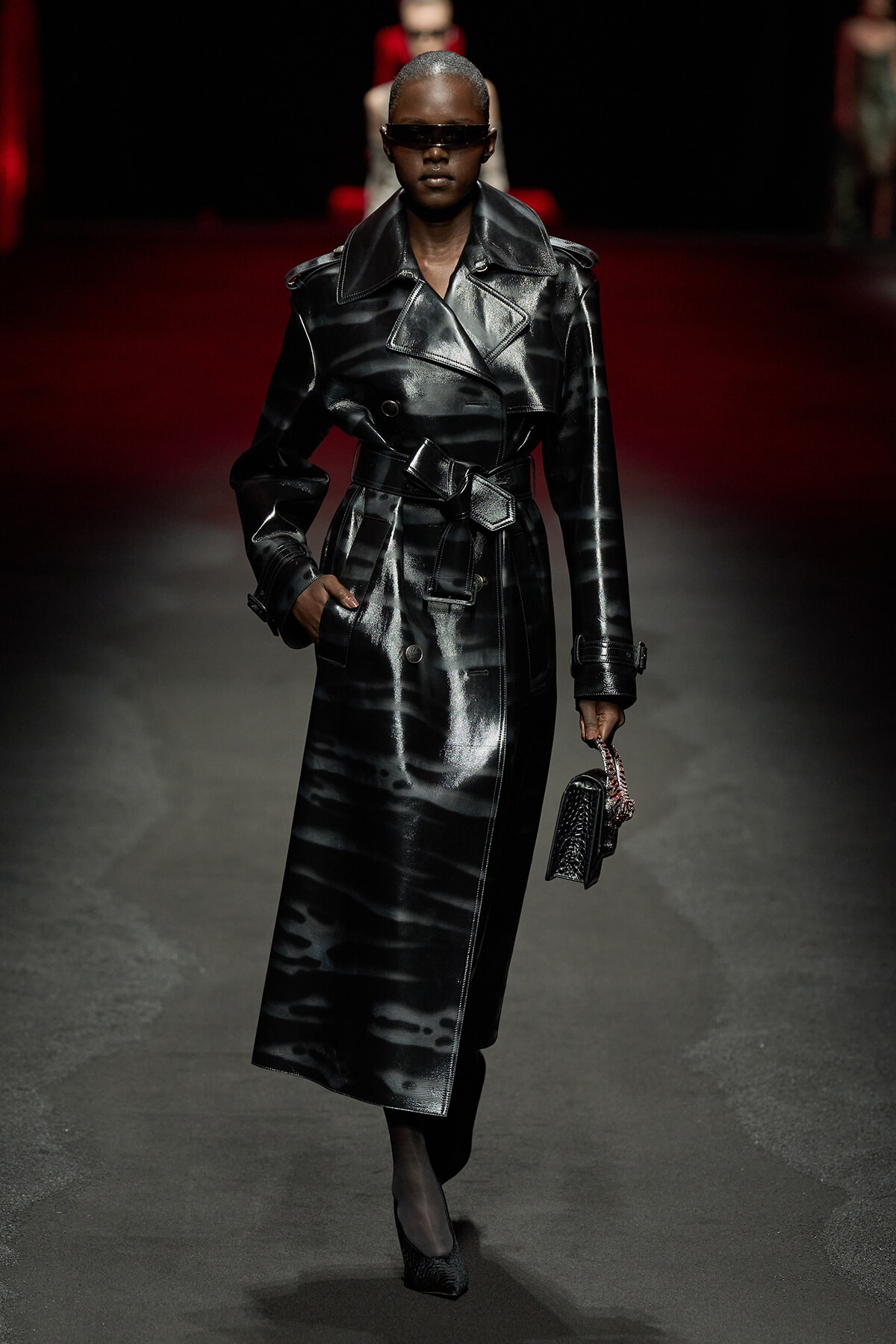 Model wearing a glossy black trench coat with a belt and oversized collar, walking a runway in dark sunglasses while holding a small black handbag.