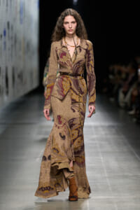 Fashion model walks the runway in a long, brown paisley-print dress with a matching belt and layered necklaces.
