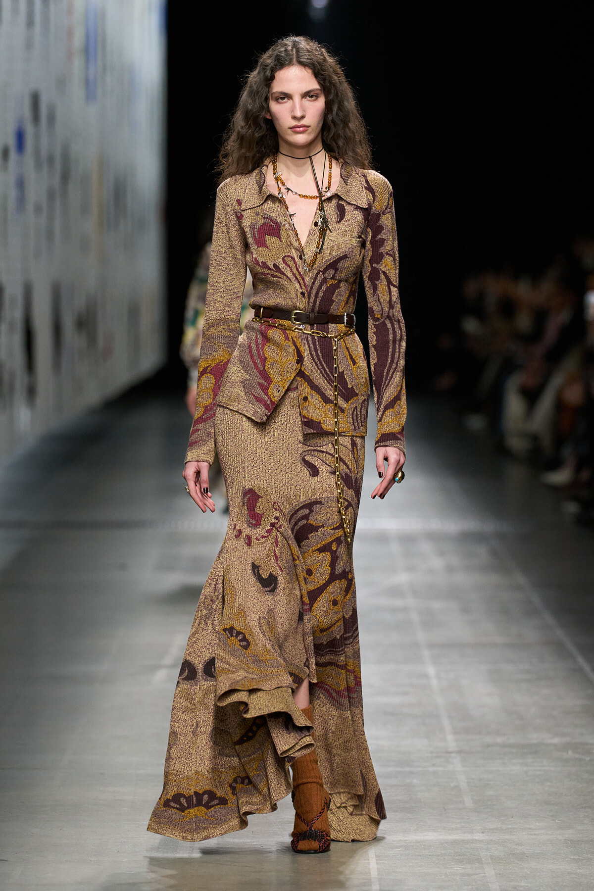 Fashion model walks the runway in a long, brown paisley-print dress with a matching belt and layered necklaces.