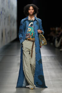 Model on a runway wearing a long blue denim coat with prints, a colorful sweater, yellow gingham pants, a brown belt, and a patterned shoulder bag.