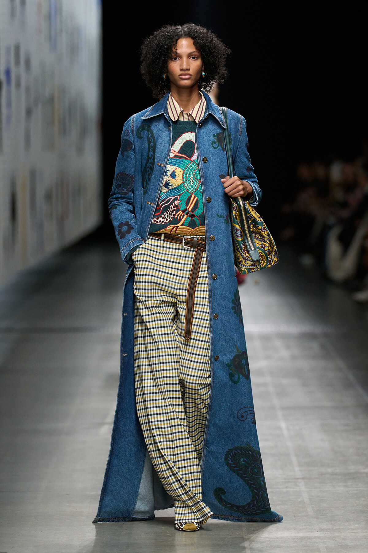 Model on a runway wearing a long blue denim coat with prints, a colorful sweater, yellow gingham pants, a brown belt, and a patterned shoulder bag.