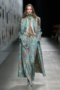 Model walks a runway in a long gray floral suit with a matching blouse and scarf, elaborate earrings visible.