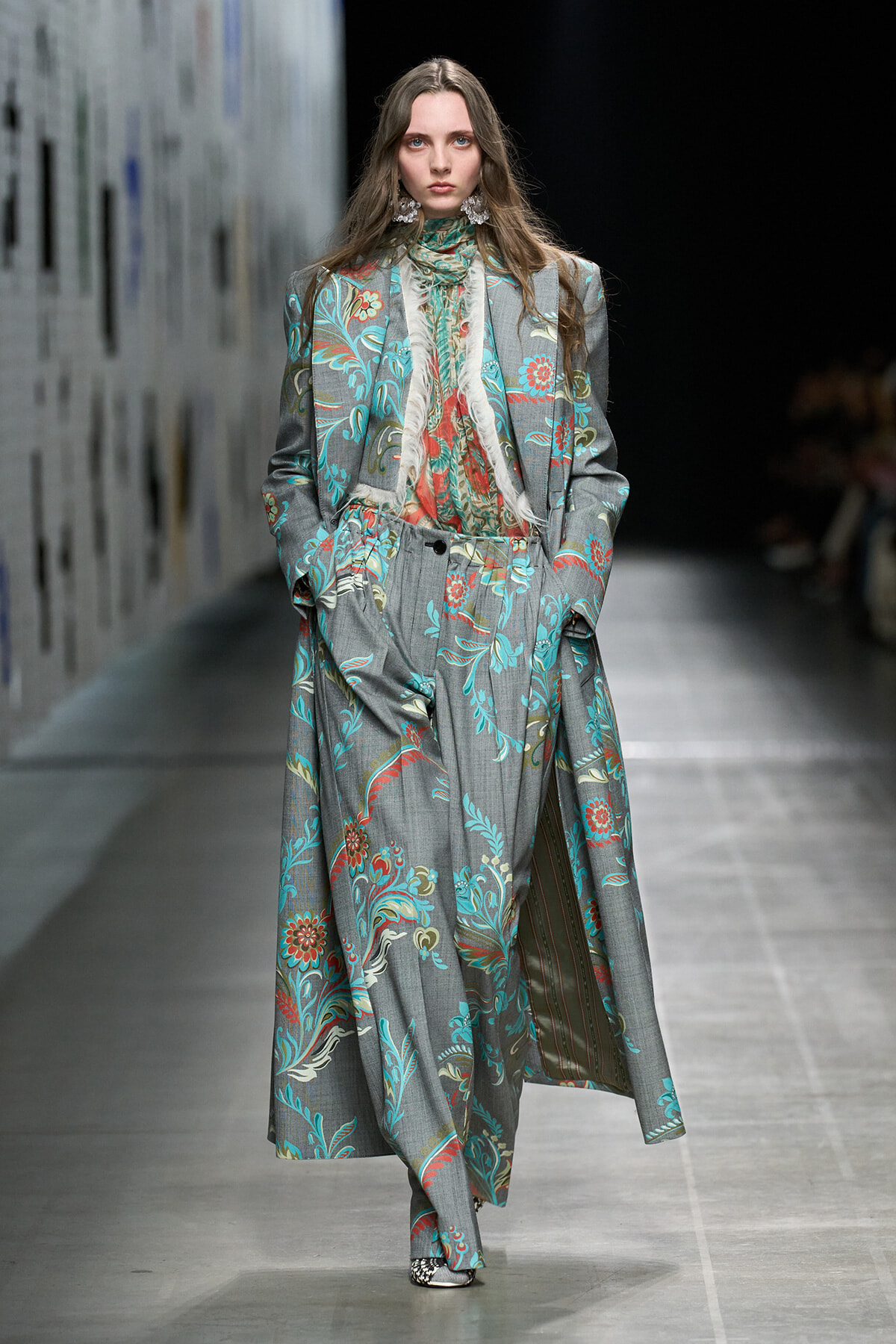 Model walks a runway in a long gray floral suit with a matching blouse and scarf, elaborate earrings visible.