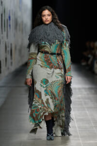 Model walks the runway in a floral-toned knit dress with a gray fringe cape and belt, accessorized with chains and patterned socks/heels.