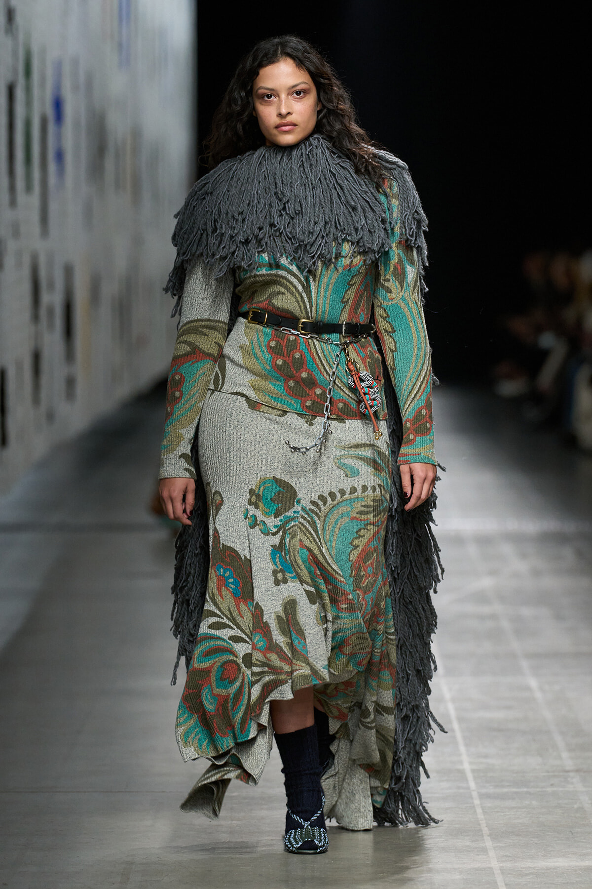 Model walks the runway in a floral-toned knit dress with a gray fringe cape and belt, accessorized with chains and patterned socks/heels.