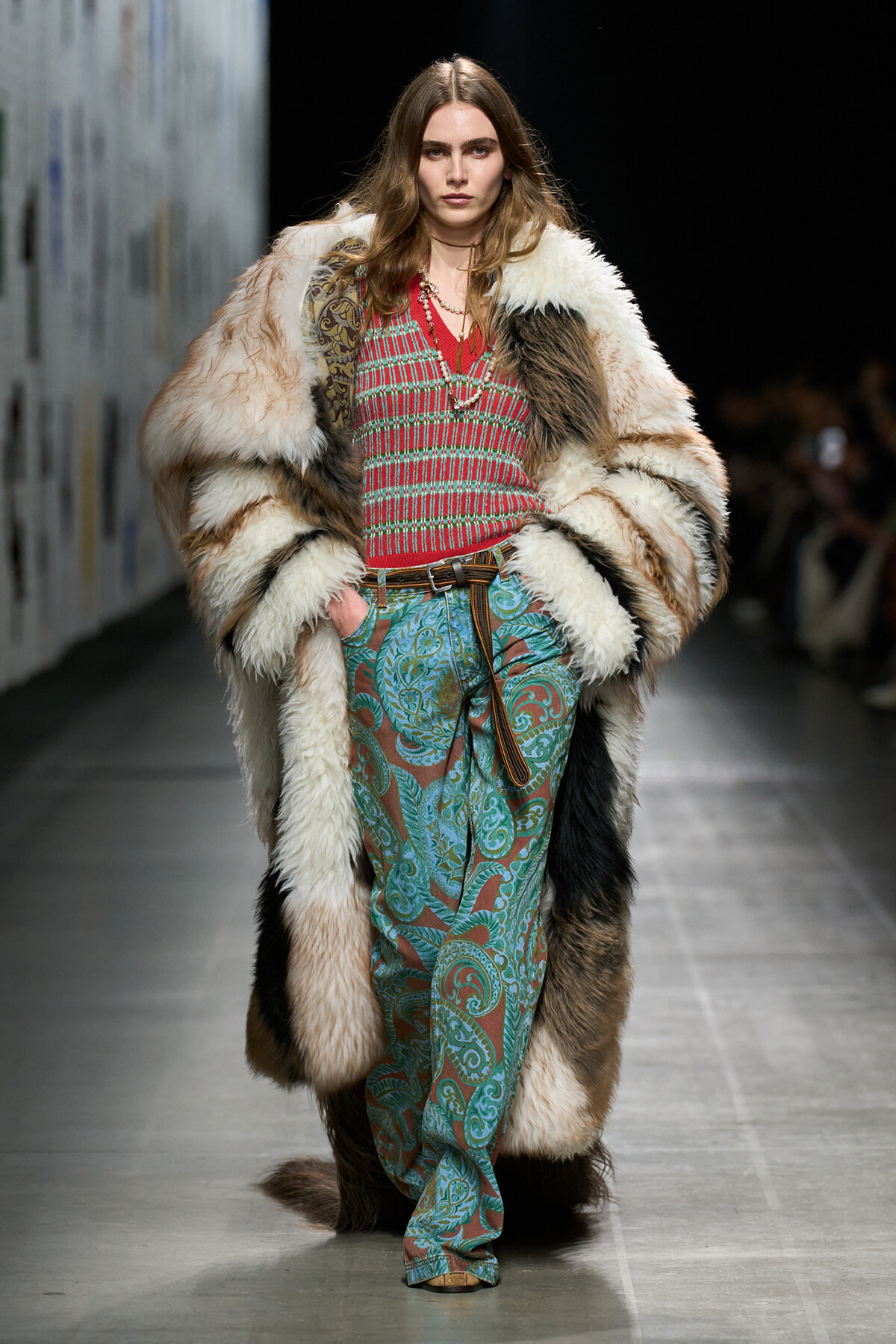Model walking a fashion runway in an oversized multicolored fur coat over a red striped knit top and turquoise paisley pants, with layered necklaces.