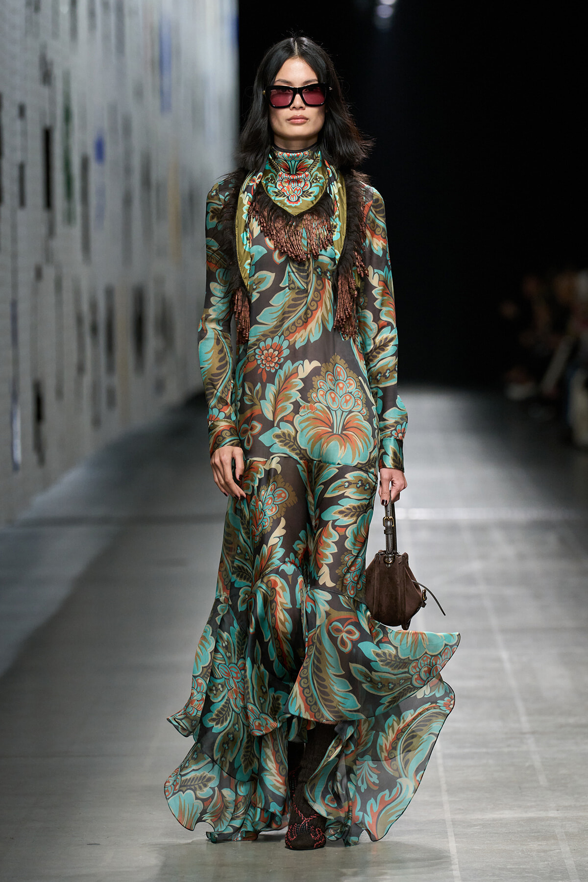 Model walking a runway in a long teal-brown floral print gown with fringe scarf and dark sunglasses, carrying a brown handbag on her left side.