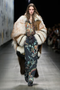 Model walks a runway wearing a oversized fur coat over a sheer floral-print denim jumpsuit, accessorized with a belt and layered necklaces.