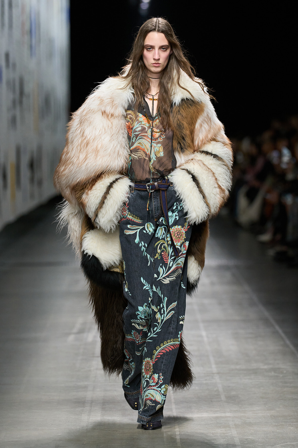 Model walks a runway wearing a oversized fur coat over a sheer floral-print denim jumpsuit, accessorized with a belt and layered necklaces.