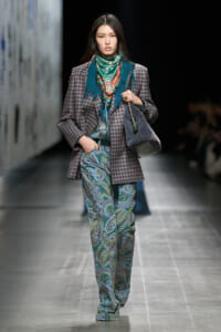 Female model walking a runway in a plaid blazer, turquoise scarf, and paisley-print trousers, carrying a gray handbag.