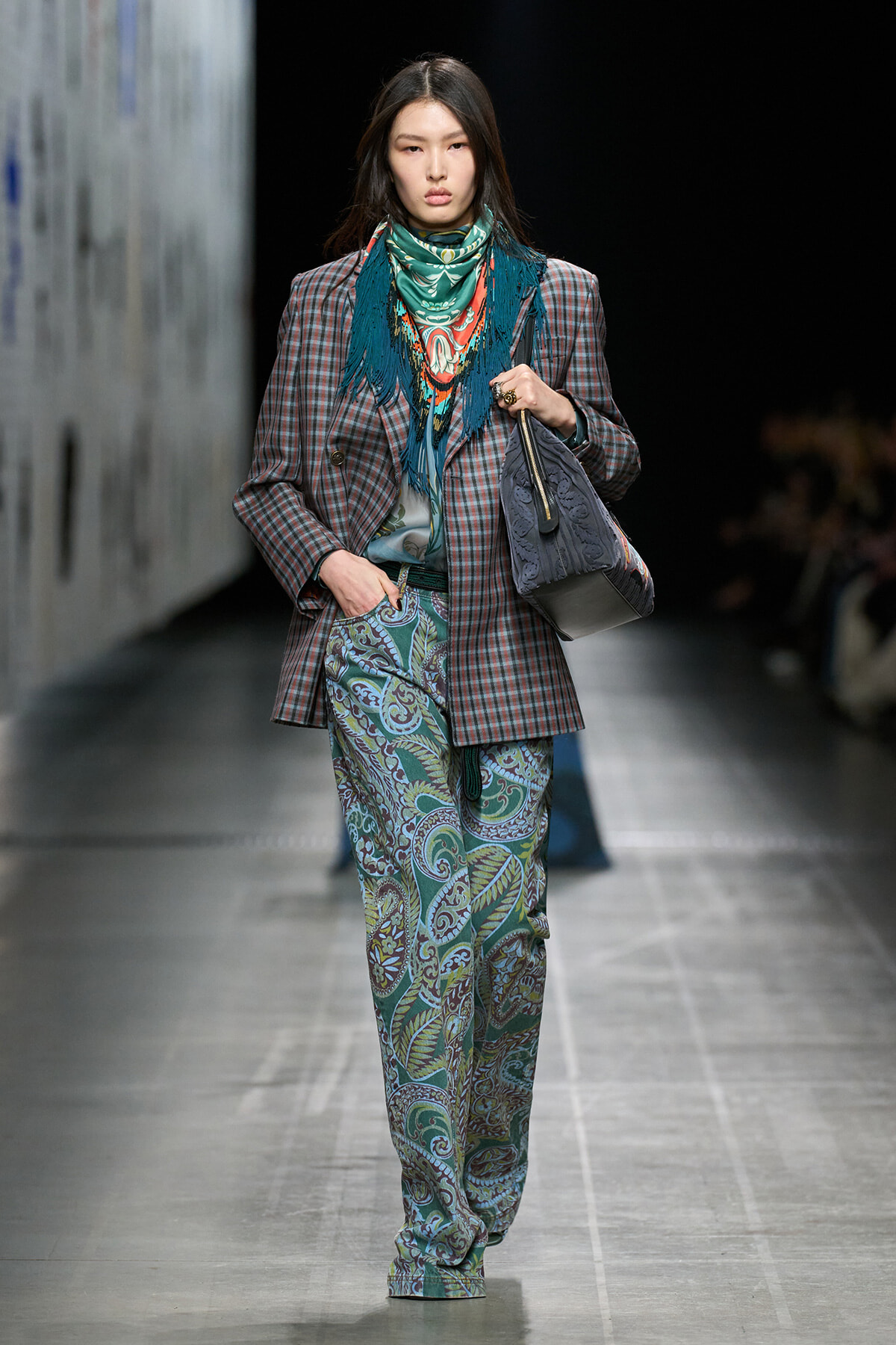 Female model walking a runway in a plaid blazer, turquoise scarf, and paisley-print trousers, carrying a gray handbag.