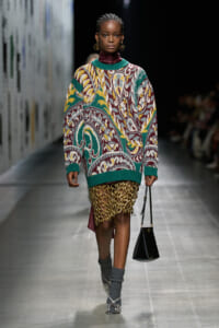 Model walking a runway in a bold, multicolored oversized knit sweater paired with a gold chain-fringe skirt and gray patterned socks with heeled shoes, carrying a black handbag.