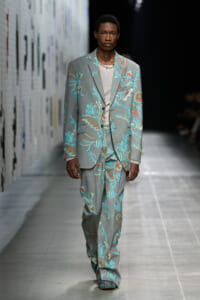 Male model walks the runway in a gray floral suit with turquoise and coral embroidery, light top underneath, during a fashion show.